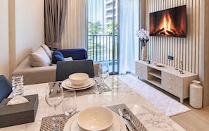 A modern living room with a sofa, a dining table set for two, a flat-screen TV mounted on a wall, and a view of the outdoors. - OZON OASIS Apartment by TVC (Choeng Thale)