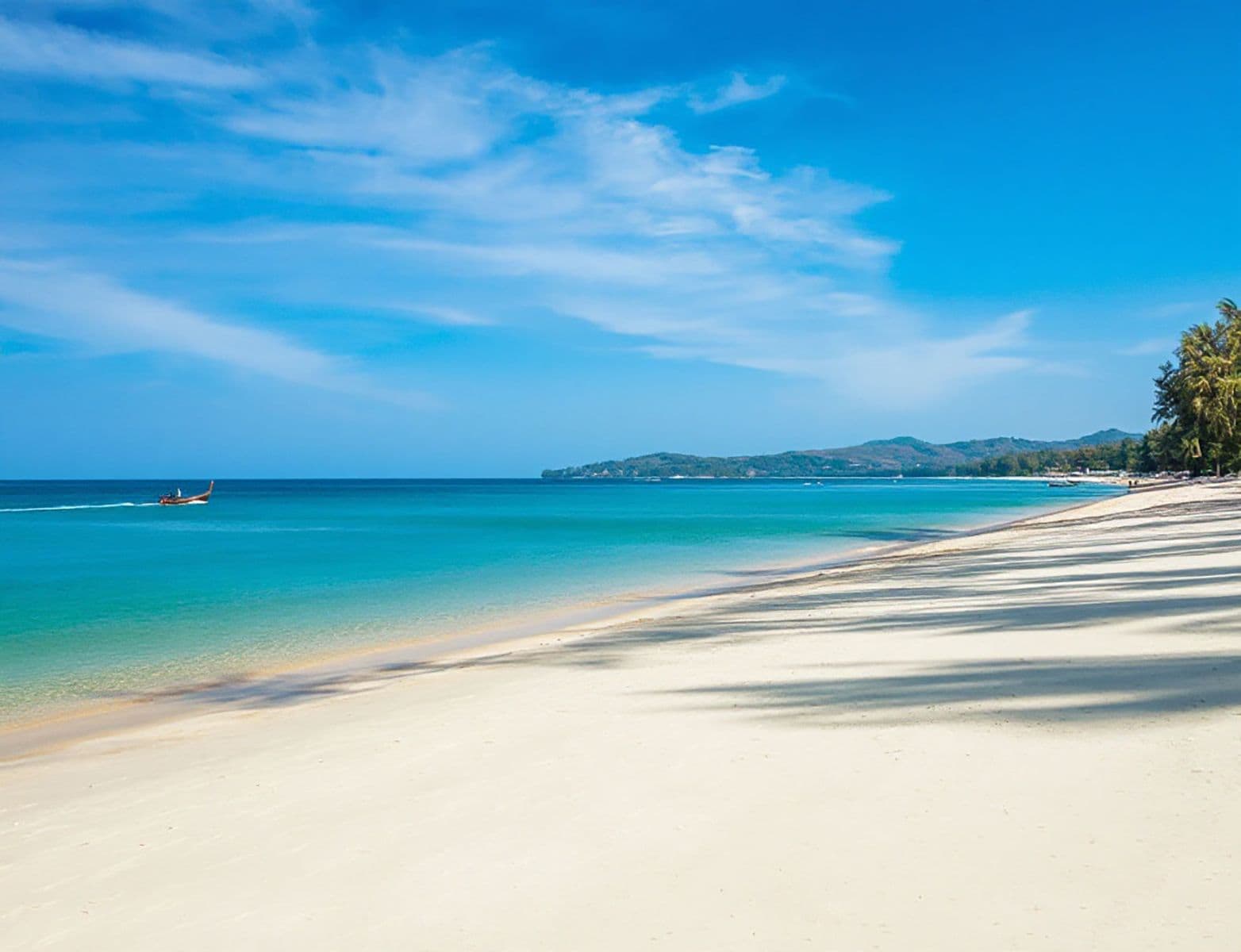 Une plage à l’eau d’un bleu cristallin, un bateau à l’horizon et une chaîne de montagnes en arrière-plan.