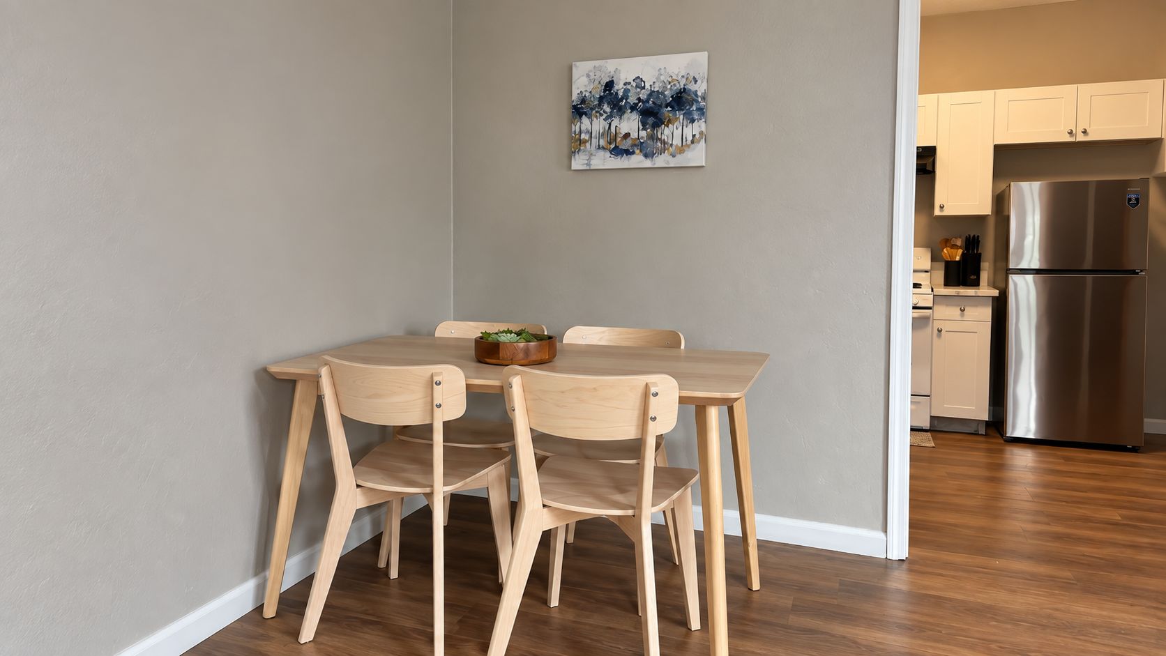 A dining area with a wooden table and chairs, a painting on the wall, and a kitchen with a stainless steel refrigerator in the background.