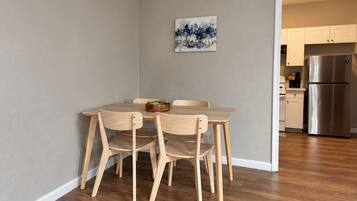 A dining area with a wooden table and chairs, a painting on the wall, and a kitchen with a stainless steel refrigerator in the background.