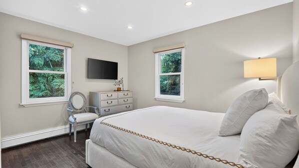 Un dormitorio con cama, cómoda, silla y dos ventanas con vistas a zonas verdes.