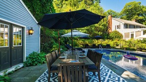 Un área de comedor al aire libre con piscina, mesas y sillas de madera, y sombrillas.