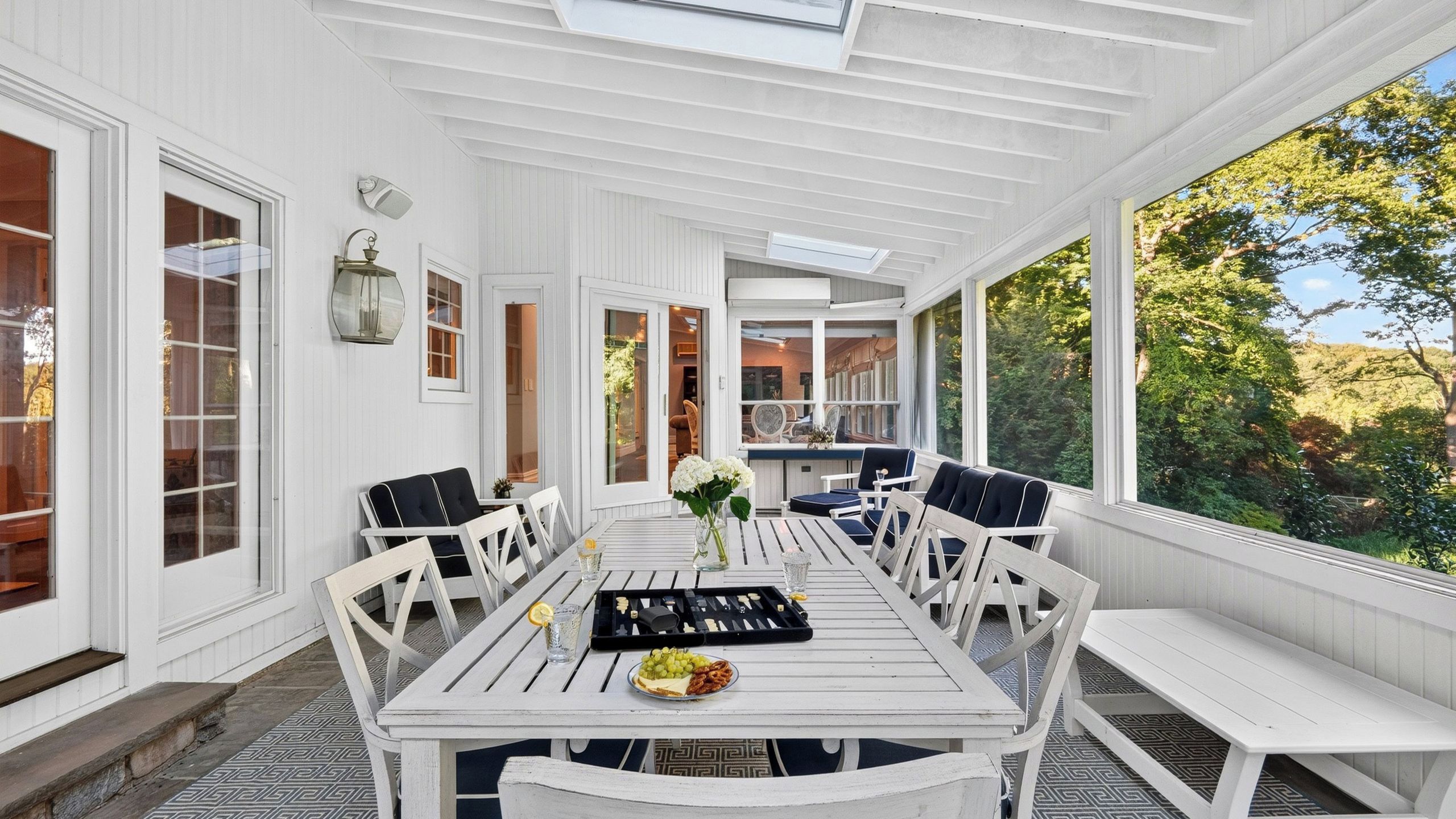 A covered outdoor dining area with a table set for four, surrounded by white chairs, and large windows offering a view of the outdoors.