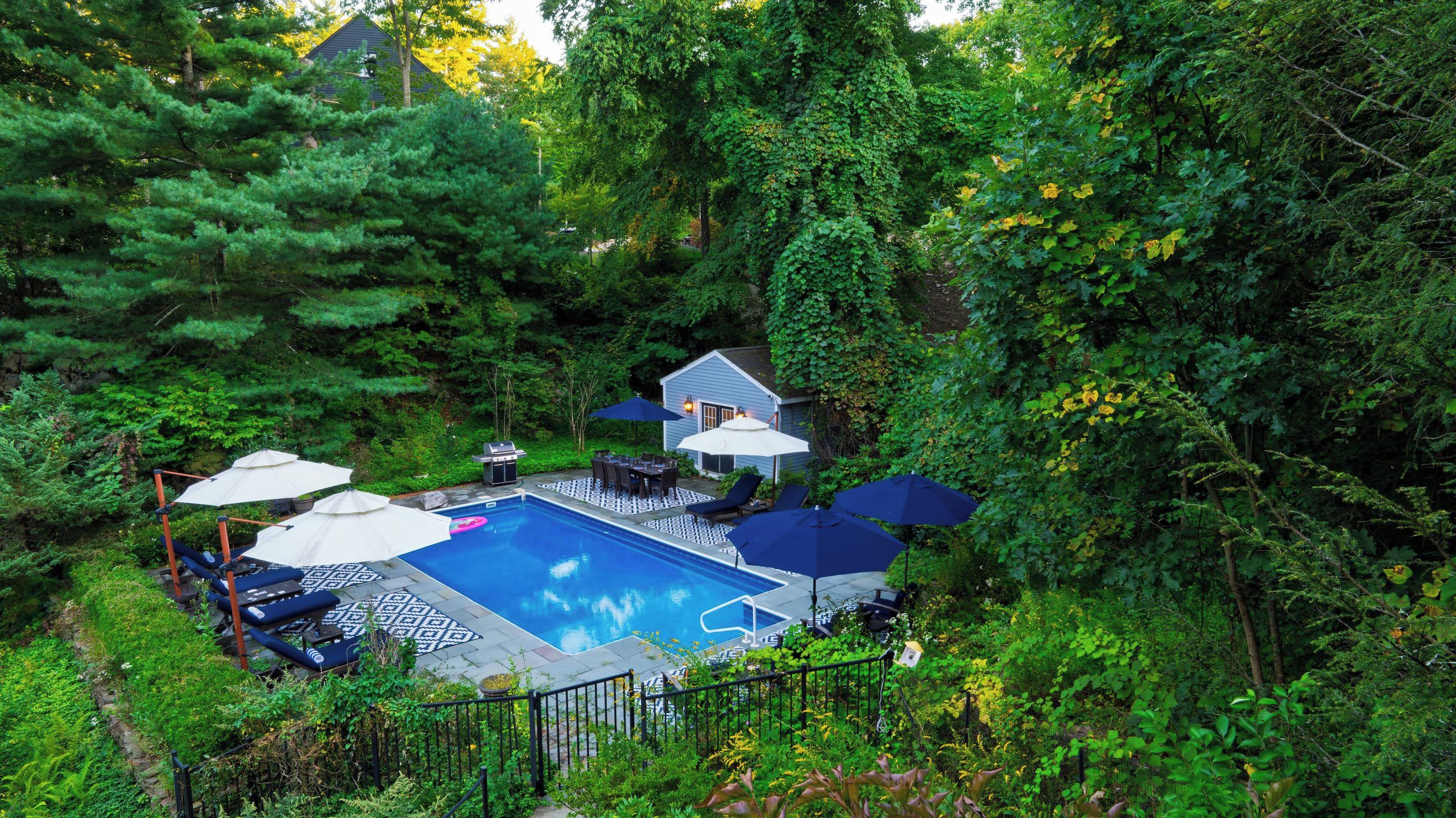 A backyard with a swimming pool, surrounded by lush greenery and a small house.