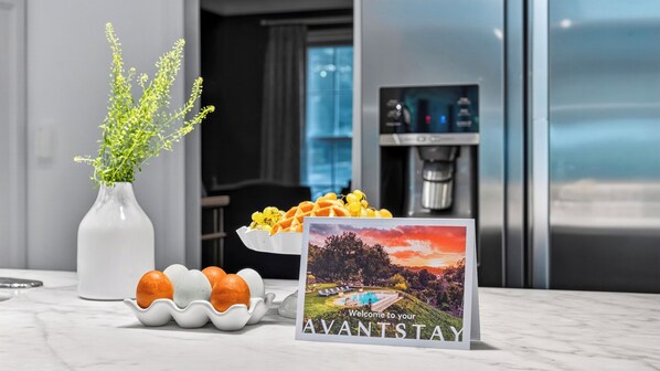 A kitchen counter with a white vase of green plants, a bowl of fruit, and a welcome sign.