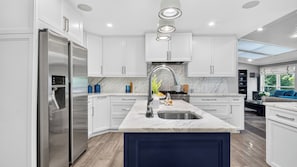 A modern kitchen with white cabinetry, a stainless steel refrigerator, a marble countertop, and a built-in island with a sink.