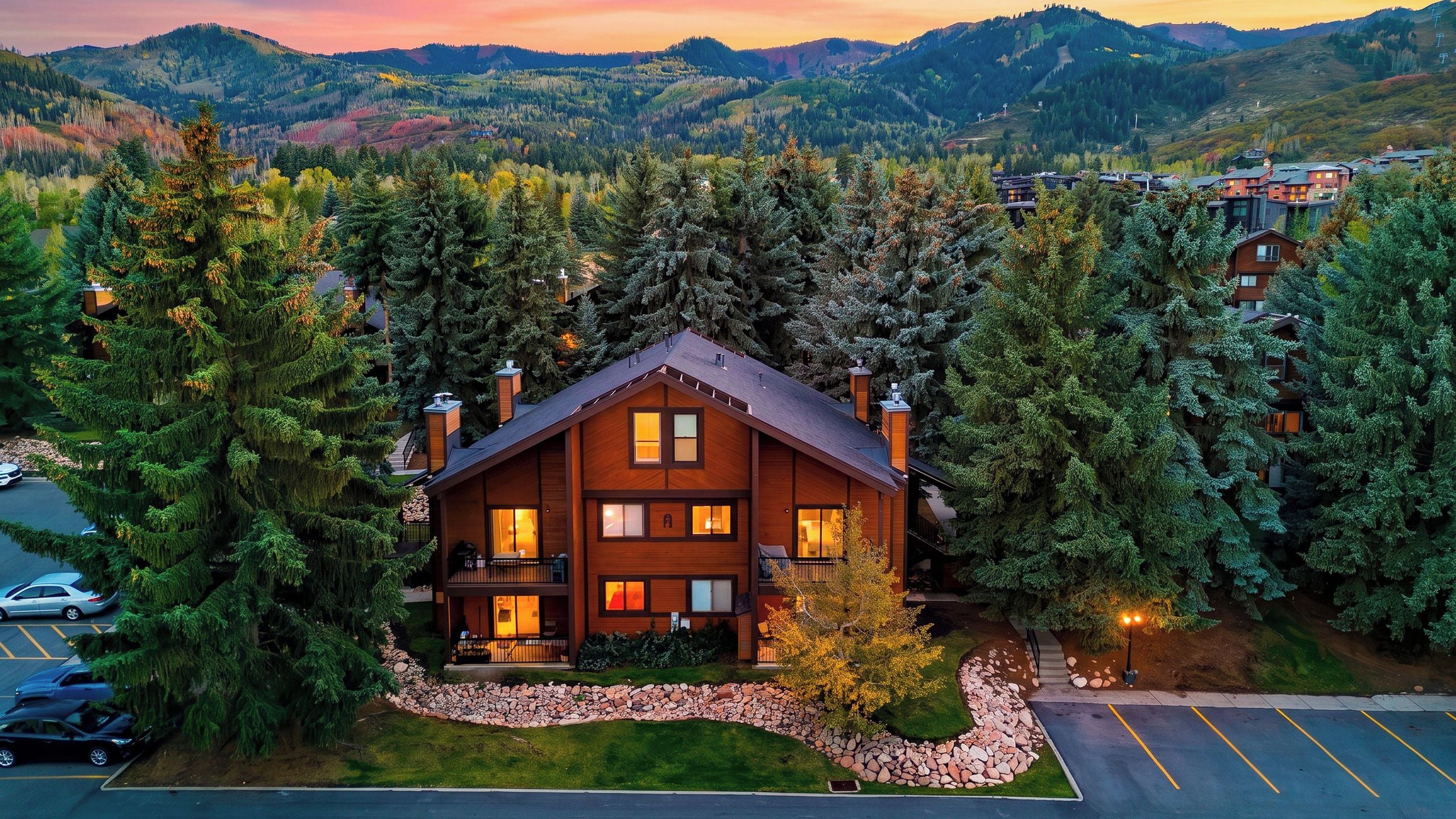 A two-story lodge with a stone foundation, surrounded by dense forest, and a parking area in the foreground.