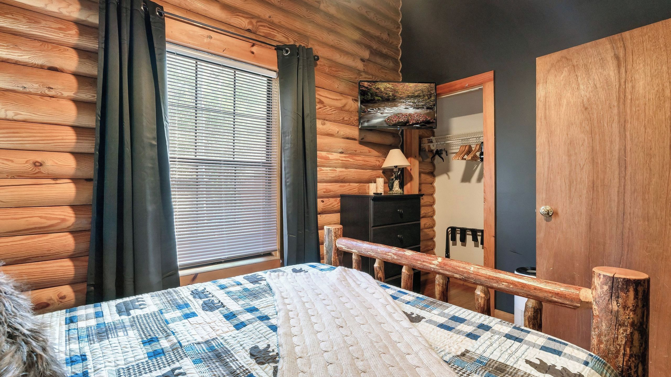 A log cabin bedroom with a bed, a dresser, a mirror, and a window with blinds.