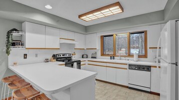 Una cocina moderna con armarios blancos, una isla central, horno empotrado, lavavajillas y ventana con vista a árboles.