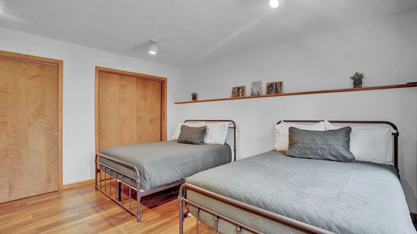 Quarto com duas camas de solteiro, soalho de madeira, guarda-roupa de madeira e prateleira com quadros.