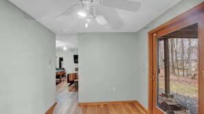 Corredor com ventilador de teto, área de refeições com mesa e cadeiras, e porta que dá para área externa com árvores.