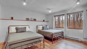 Ein Schlafzimmer mit zwei Betten mit Metallgestell, Holzboden, einem Fenster mit Blick auf Bäume und einer Ablage mit Dekoration.