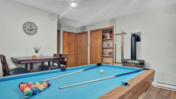 Una mesa de billar con bolas, un reloj y una estantería en una habitación con pisos de madera y paredes blancas.