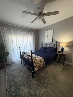 Ein Schlafzimmer mit einem Bett, einem Deckenventilator, einem Nachttisch mit Lampe, einer Topfpflanze und einem Fenster mit Vorhängen.
