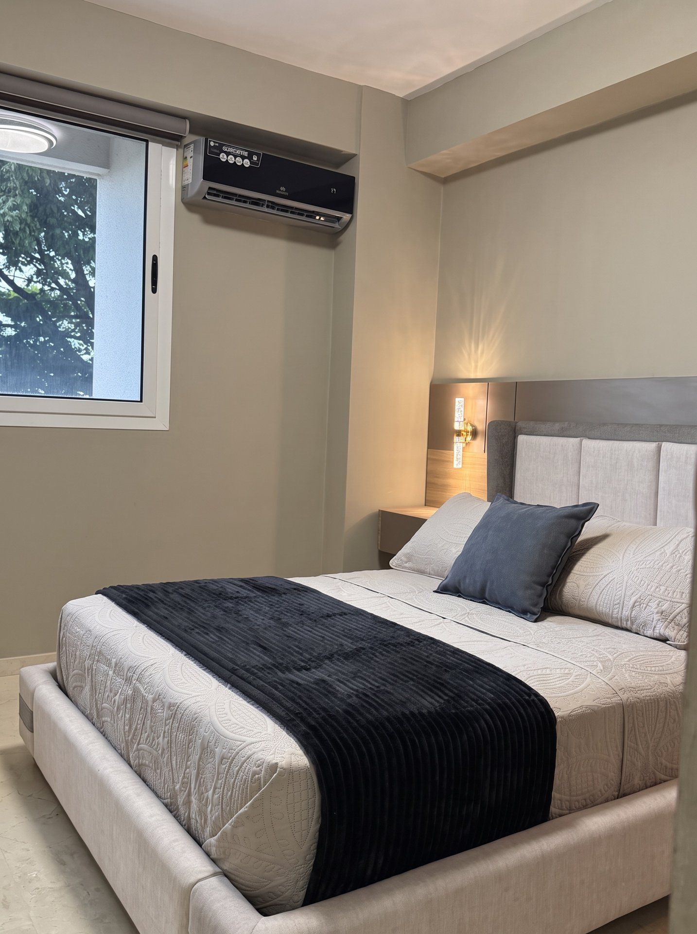 Une chambre à coucher comprenant un lit, une fenêtre, un appareil de climatisation et un luminaire fixé au mur.