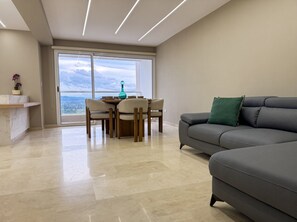 A modern living room with a grey sofa, a dining table with chairs, and a view through a large window.