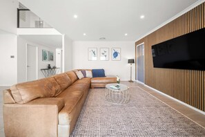 A modern living room with a large sectional sofa, a glass coffee table, and a flat-screen TV mounted on a wooden panel wall. - Sunset Cove - Bribie Island Waterfront Retreat (Bribie Island Region)