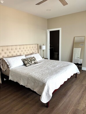 A bedroom with a tufted headboard, a bed with a white quilt, a nightstand with a lamp, and a mirror on the wall. -  New luxurious beachfront home with saltwater pool (Milton)