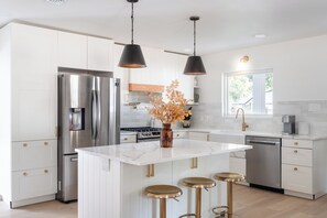 A modern kitchen with a central island, stainless steel appliances, pendant lighting, and a window providing natural light. - Luxe 4BR/3BA • Heated Pool + Gym, 5 Min Downtown (Austin)