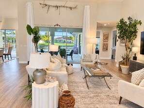 A spacious living room with a large window, a dining area, and a view of a house and palm trees. - Perfect 4-bedroom house with AC in charming Cape Coral (Cape Coral)