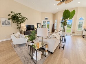 Amplia sala de estar con sofá, sillones, una mesa de centro y televisor. Hay plantas, un ventanal grande y un ventilador de techo.