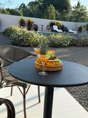 Ein runder Tisch mit einem geflochtenen Obstkorb, der eine Ananas, einen Apfel und eine Orange enthält, sowie ein Glas Orangensaft.