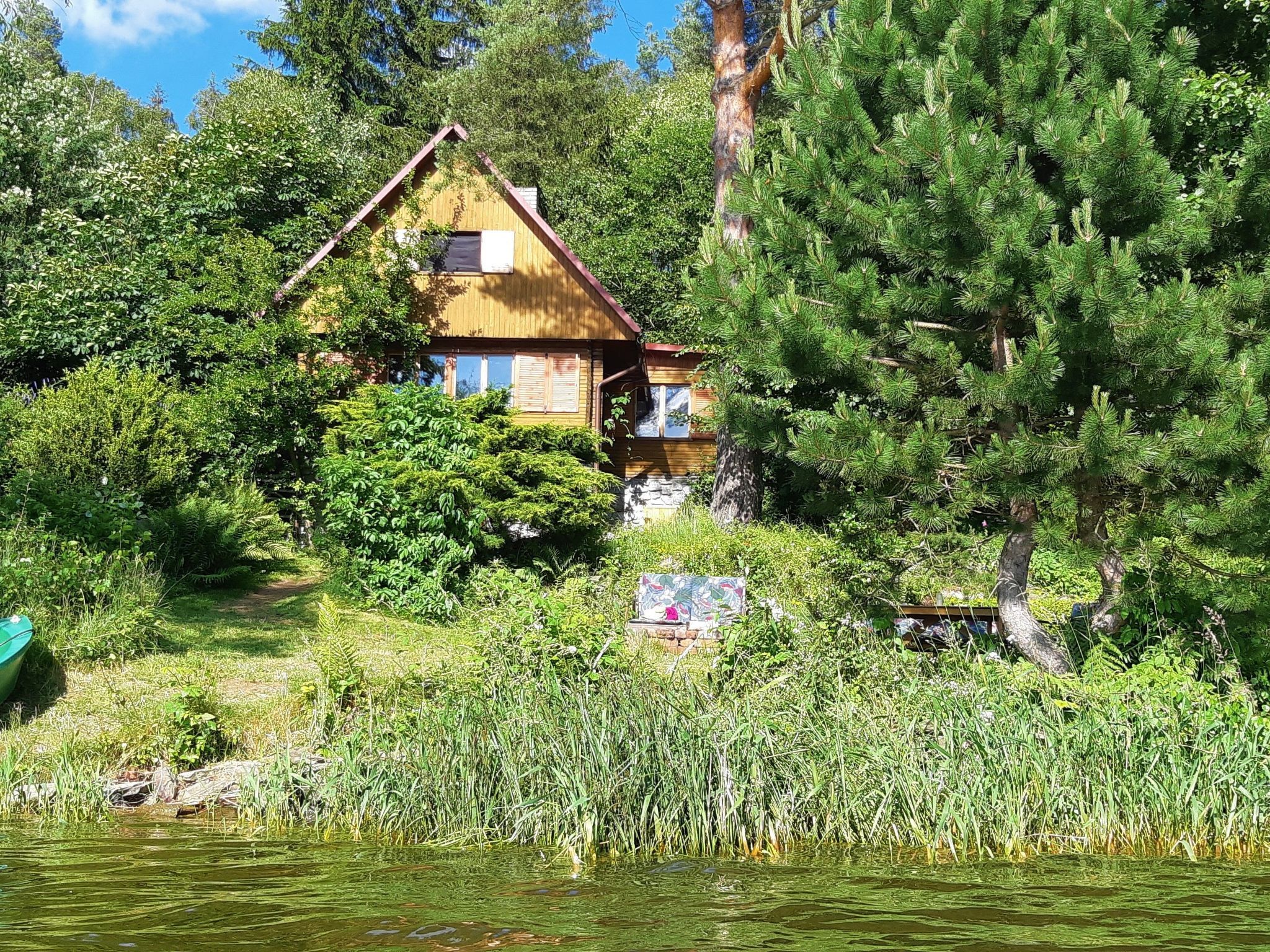 A wooden cabin with a sloped roof, surrounded by dense greenery and a body of water.