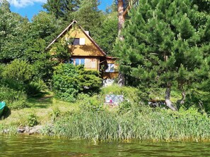 Eine Holzhütte mit geneigtem Dach, umgeben von dichtem Grün und einem Gewässer.