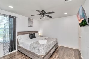Une chambre à coucher avec un lit, un ventilateur de plafond, un téléviseur fixé au mur et une fenêtre avec des rideaux.