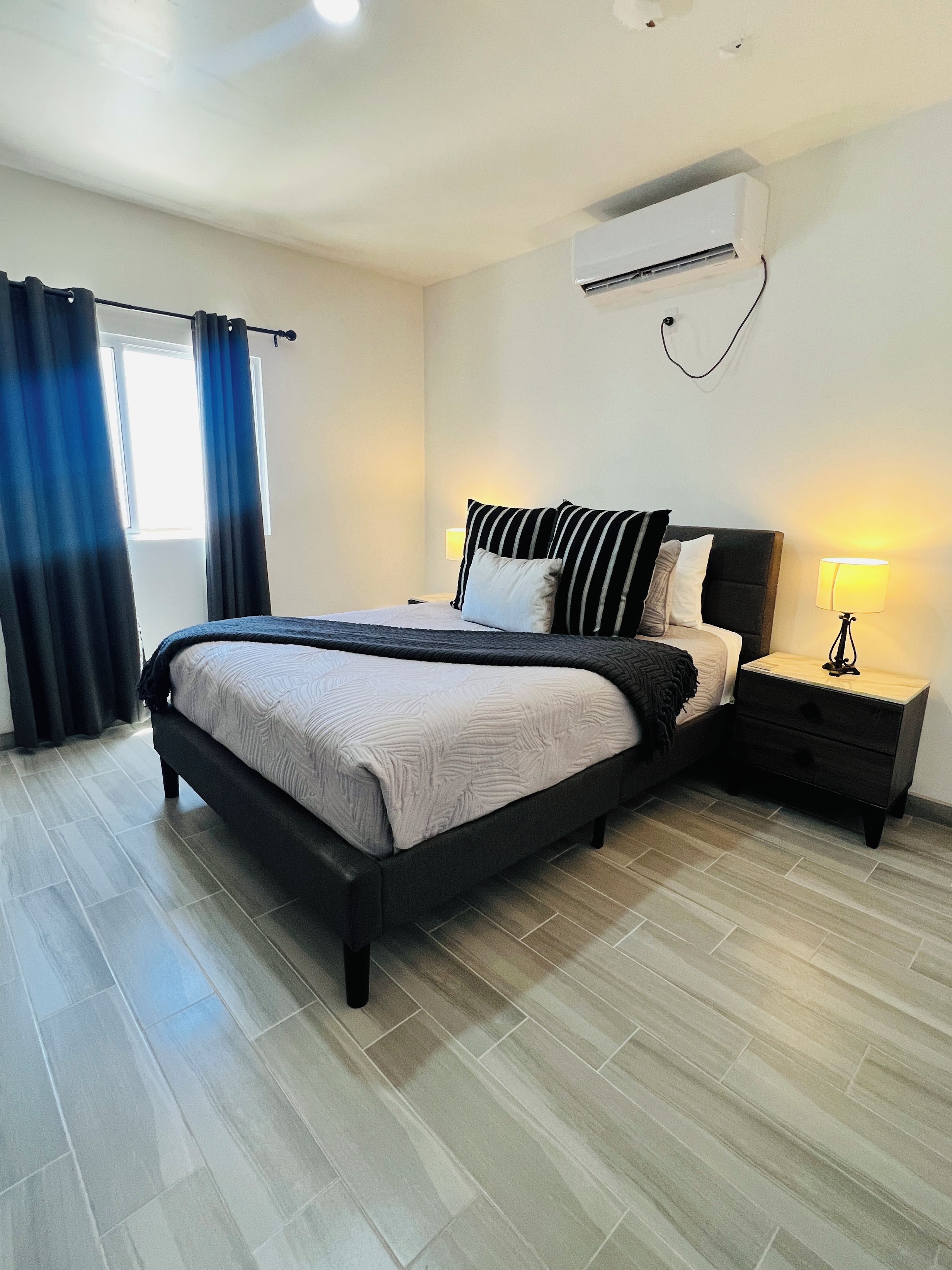 Een moderne slaapkamer met een bed, nachtkastjes, een airconditioning en een raam met gordijnen.