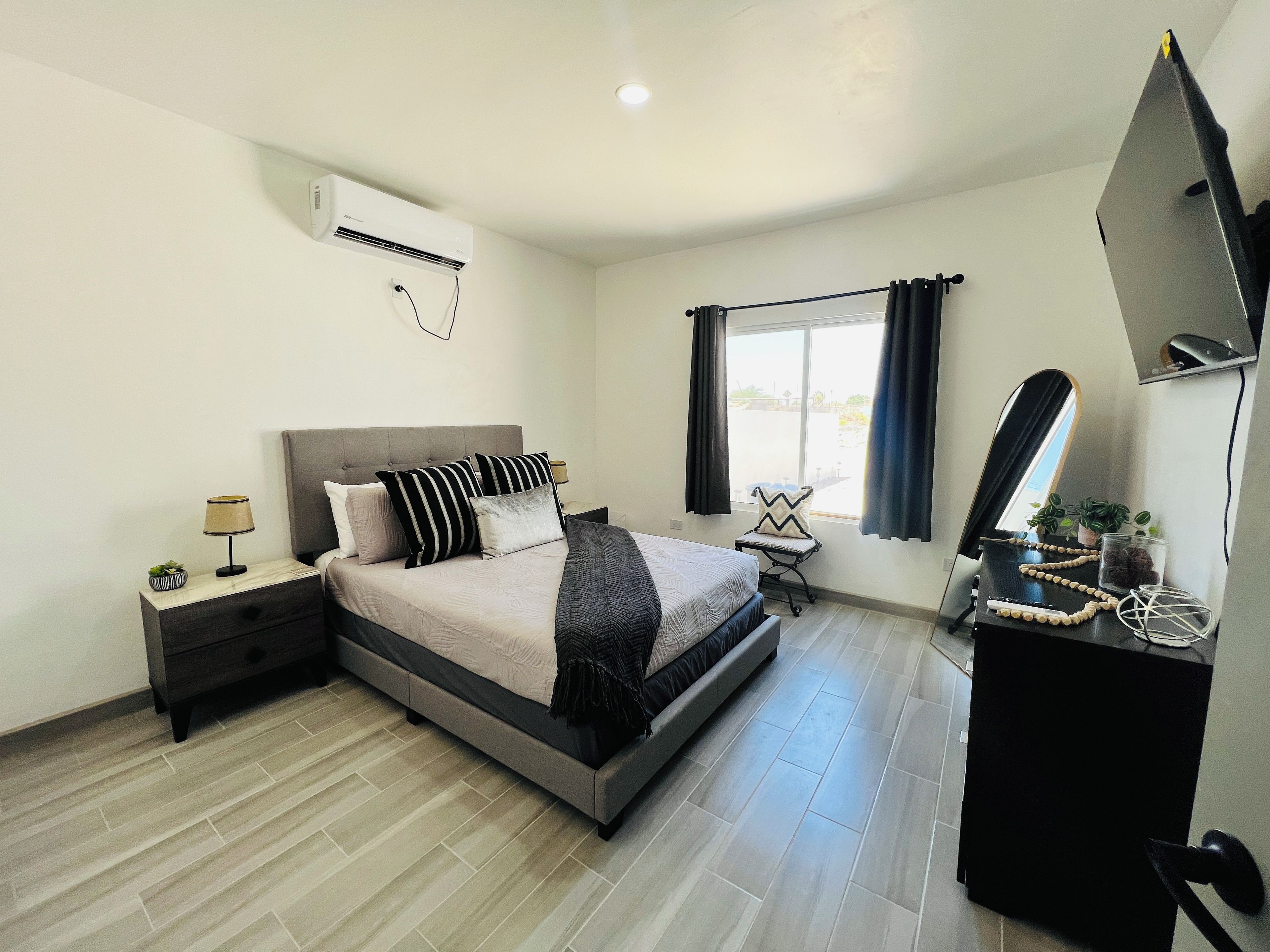 A bedroom with a bed, bedside table, lamp, air conditioning unit, window with curtains, and a desk with a mirror and plants.