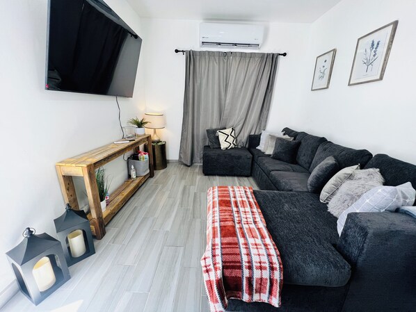A modern living room with a dark grey sofa, a wooden console table, a flat-screen TV mounted on the wall, and a grey curtain.