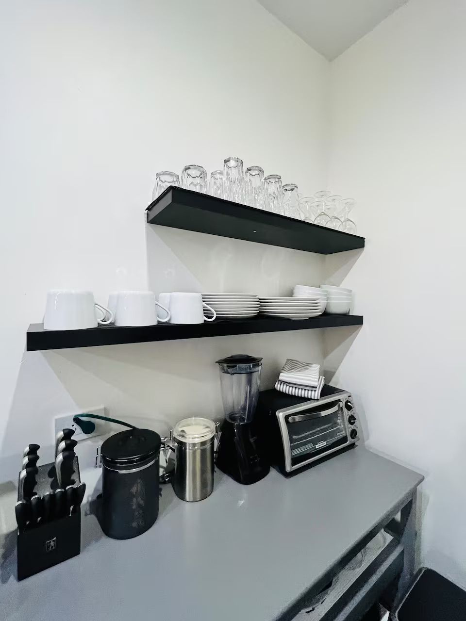 Eine Küche mit schwarzen Regalen, auf denen weiße Tassen, Teller und Gläser sowie eine Kaffeemaschine, ein Mixer und ein Toaster stehen.