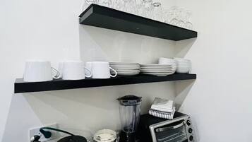 Une cuisine avec des étagères noires où sont rangées des tasses, des assiettes et des verres blancs, ainsi qu’une machine à café, un mixeur et un grille-pain.