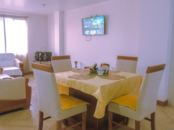 A dining area with a table set for four, a television mounted on the wall, and a window with curtains.
