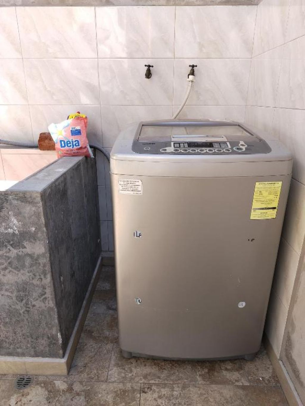 A compact, beige water heater with a control panel, located in a corner with tiled walls and a concrete floor.