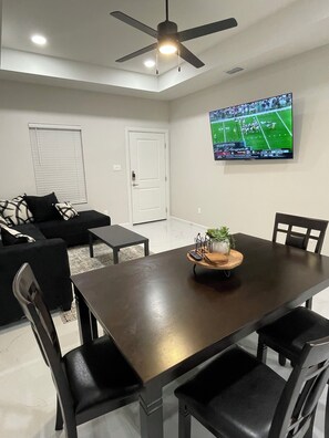 A room with a dark wooden table, black chairs, a ceiling fan, a TV on the wall, and a door. - Apartment in Weslaco, TX. 3BR / 2 BA (Weslaco)