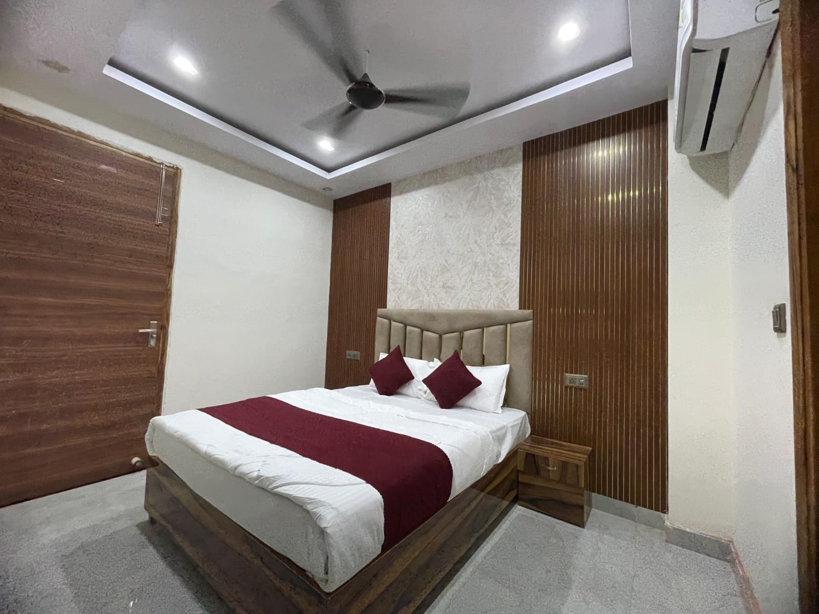 A hotel room with a bed, a ceiling fan, an air conditioning unit, and wooden paneling on the walls.
