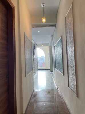 A hallway with a wooden door on the left, a light fixture overhead, and artwork on the walls. - Hotel White House  (Ghaziabad)