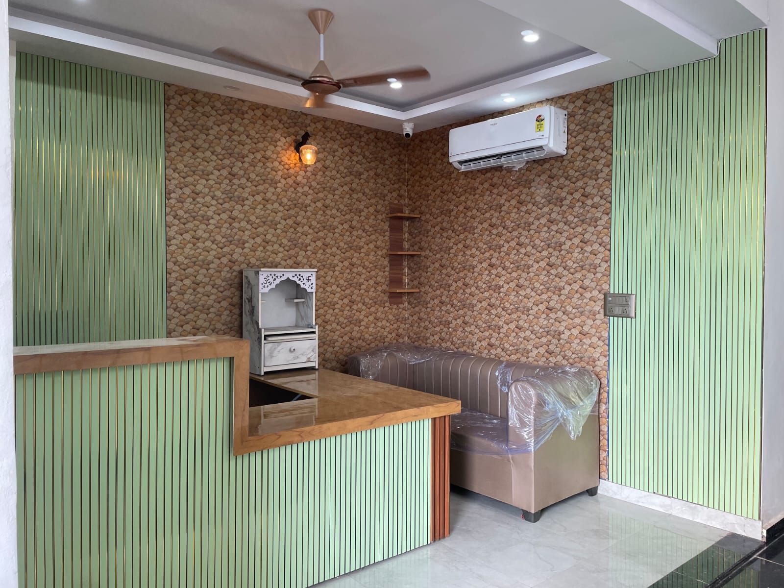 Un área de recepción con mostrador de madera, zona de estar con un banco acolchado, unidad de aire acondicionado en la pared y ventilador de techo.