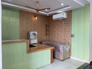 A reception area with a wooden reception desk, a seating area with a cushioned bench, a wall-mounted air conditioning unit, and a ceiling fan. - Hotel White House  (Ghaziabad)