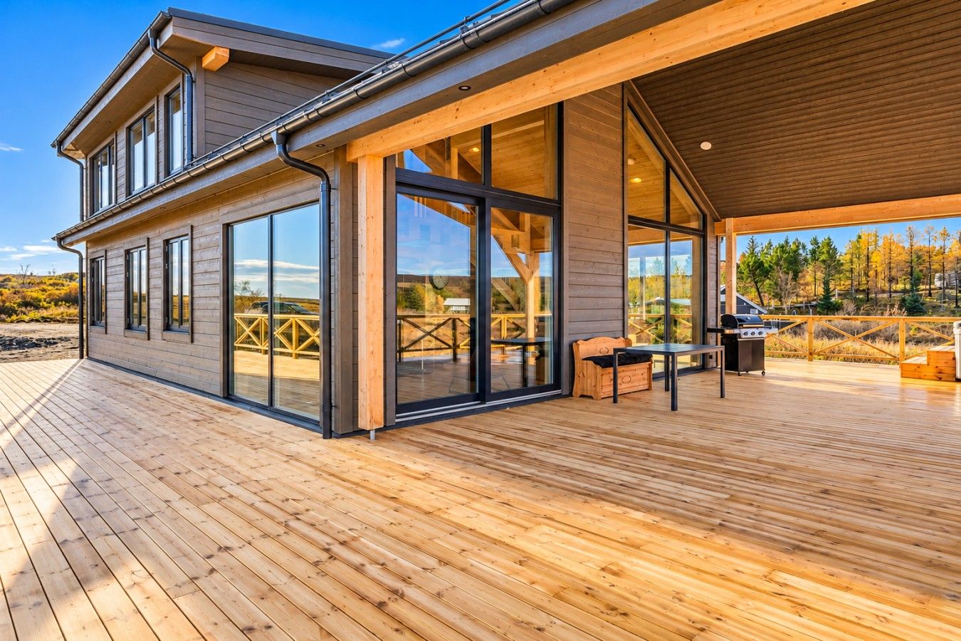 A modern wooden house with large glass windows, a deck, and outdoor furniture.