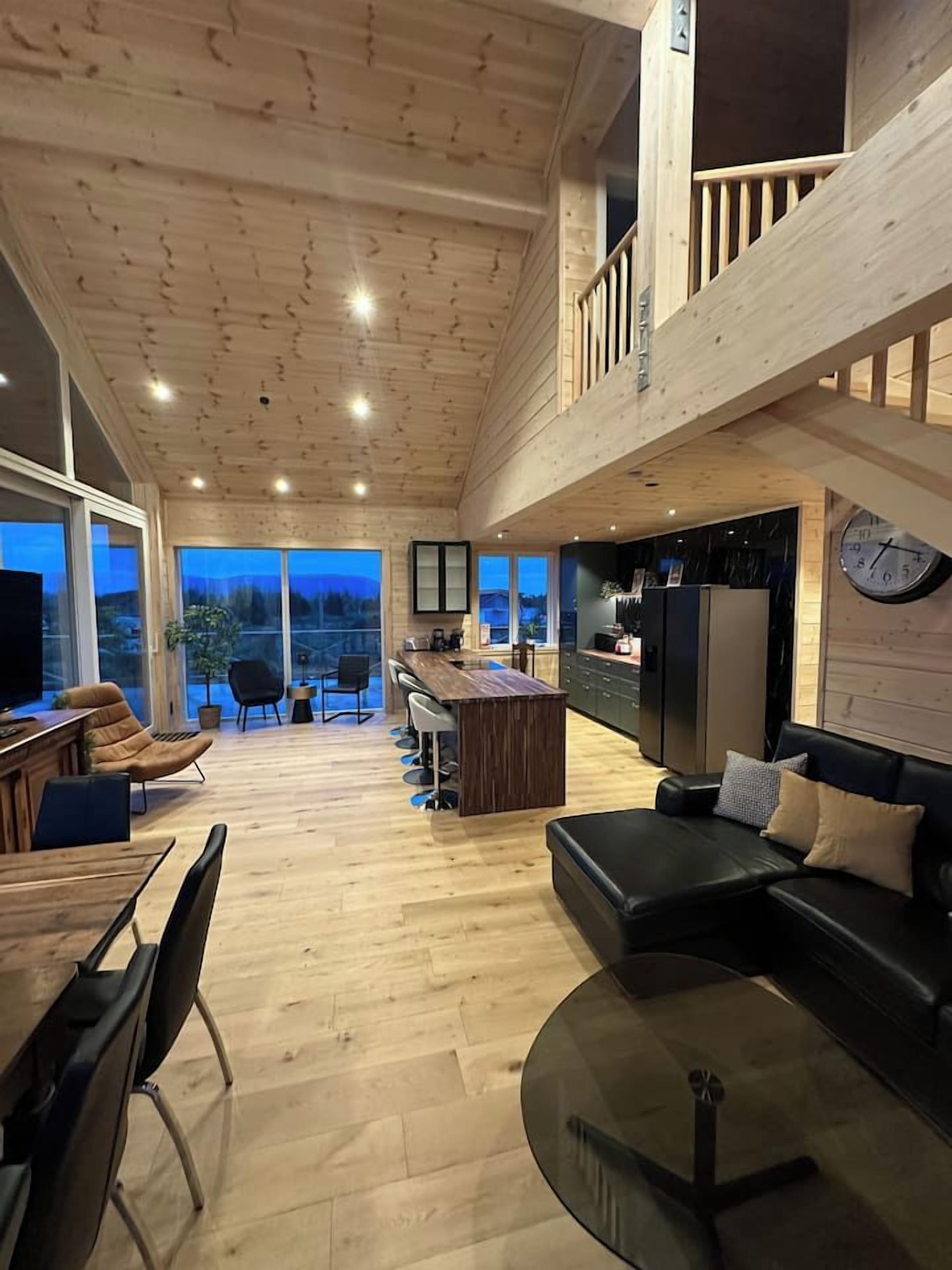 Un vaste espace de vie doté d’un haut plafond, d’un plancher en bois, d’un canapé en cuir noir, d’une table à manger avec des chaises, et d’un coin cuisine comprenant un réfrigérateur et une horloge murale.