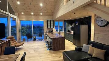 Un vaste espace de vie doté d’un haut plafond, d’un plancher en bois, d’un canapé en cuir noir, d’une table à manger avec des chaises, et d’un coin cuisine comprenant un réfrigérateur et une horloge murale.