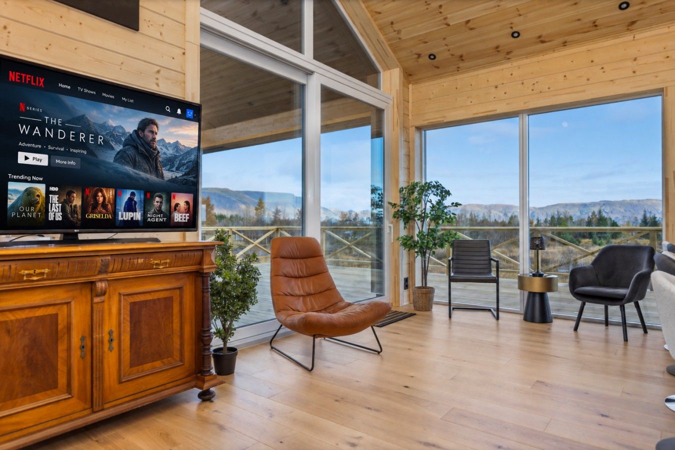 A modern living room with a large TV displaying a Netflix series, wooden furniture, and a balcony with a view of the mountains.