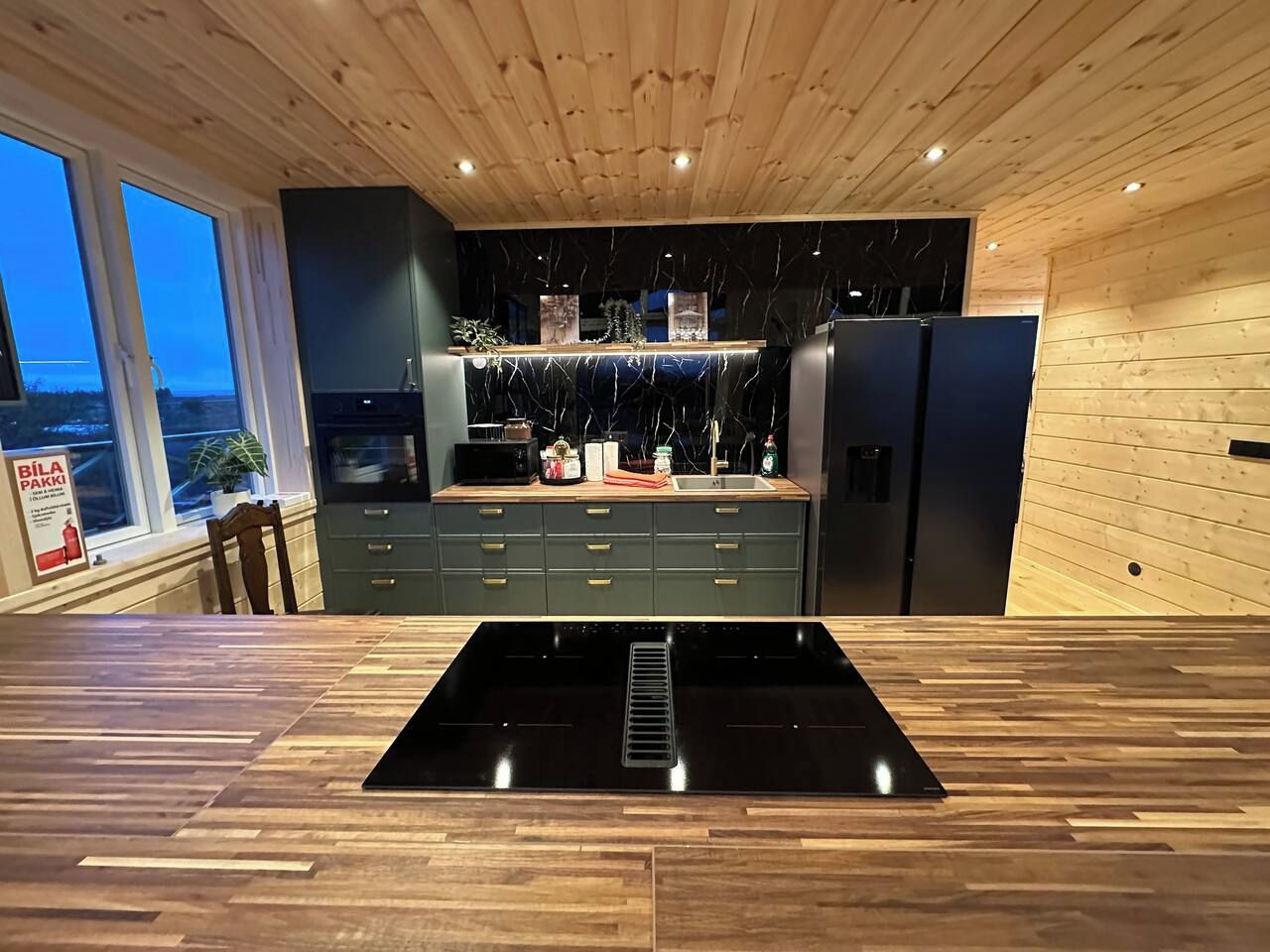 A modern kitchen with a central island, dark cabinetry, and a view through large windows.
