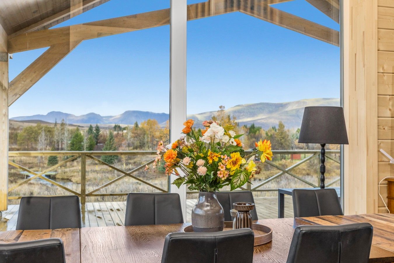 A dining area with a table set for four, a vase of flowers, and a view of mountains through large windows.