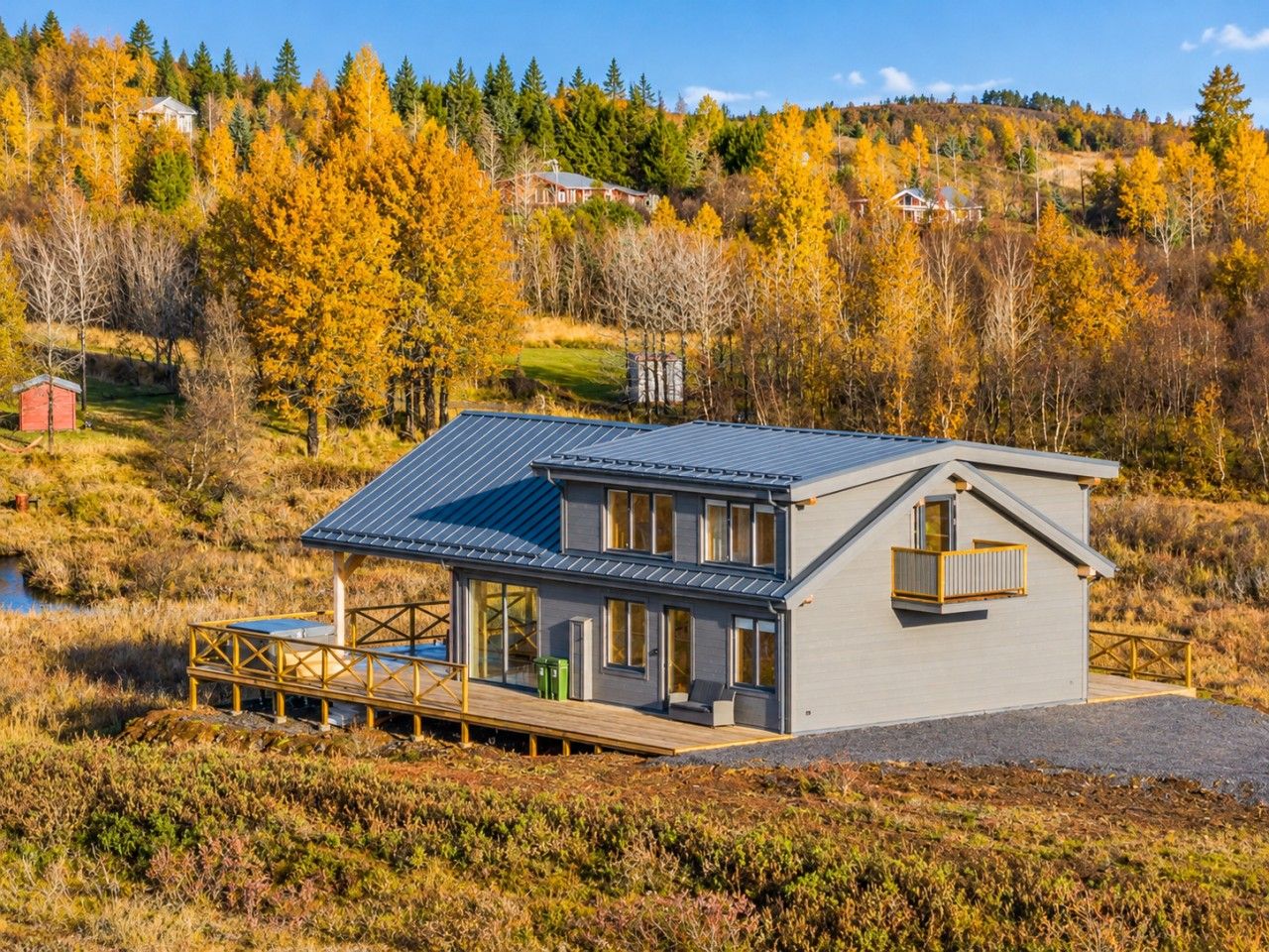 A modern two-story house with a deck, surrounded by autumn foliage and a hill in the background.