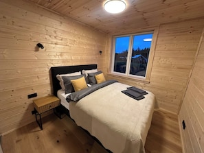 Ein Schlafzimmer mit Holzwänden, einem Bett, einem Nachttisch und einem Fenster mit Blick auf Häuser.