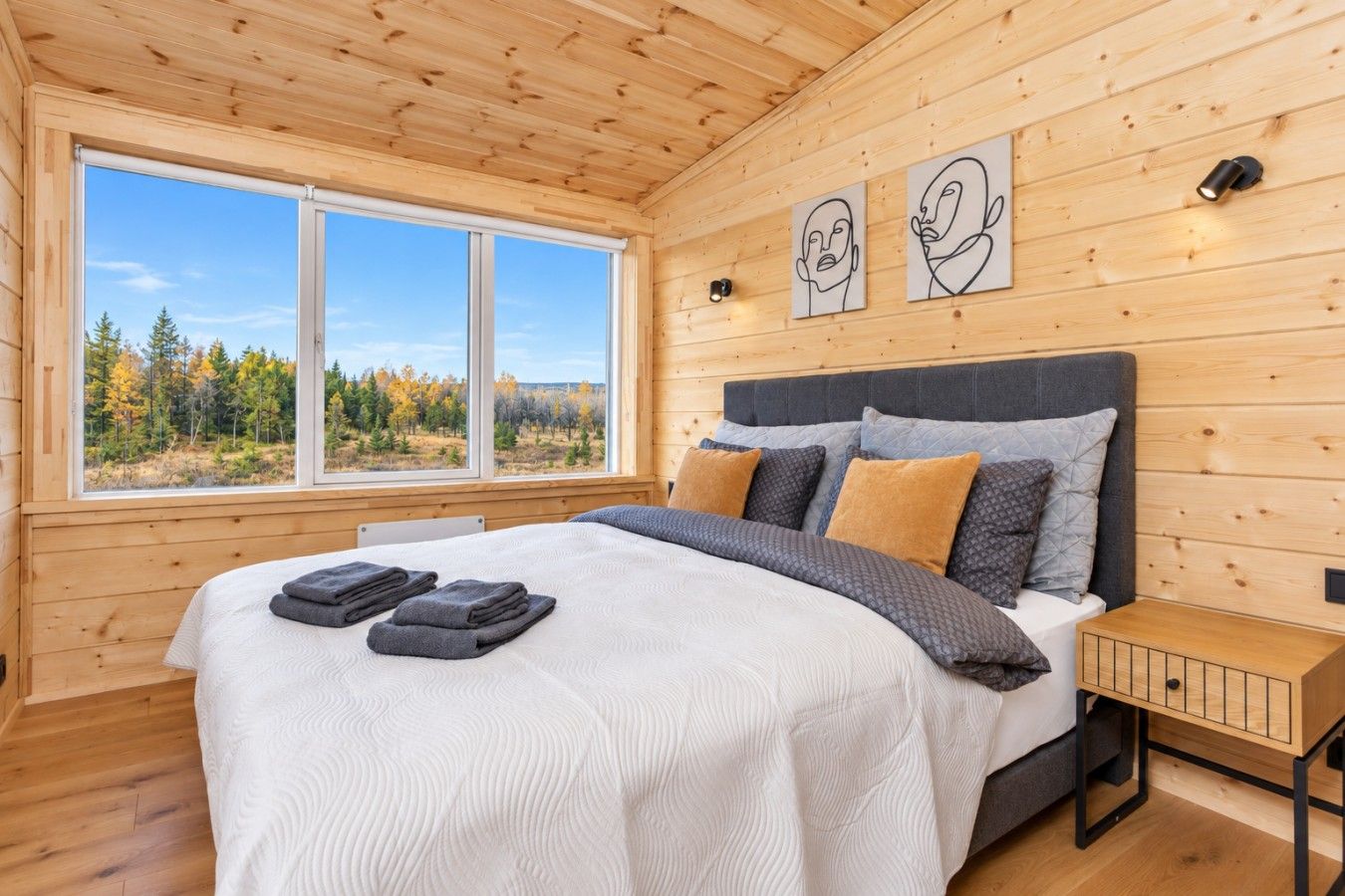 Une chambre avec un grand lit, des murs en bois, une fenêtre donnant sur les arbres et deux appliques murales.